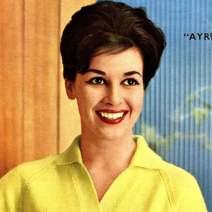 May include: A woman with dark hair styled in a bouffant wears a yellow collared sweater. She is smiling and looking to the right of the frame. The background is a blue and wood-paneled wall.
