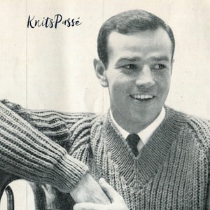 May include: A black and white photograph of a man wearing a knitted jumper with a V-neck. The jumper has a cable knit pattern. The man is smiling and has his arm resting on a surface.