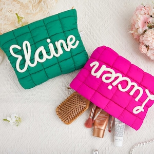 Puede incluir: Dos neceseres acolchados, uno verde esmeralda y otro rosa intenso, cada uno con un nombre blanco en cursiva. La bolsa verde dice "Elaine", y la bolsa rosa dice "Lucien". Pinceles de maquillaje, un cepillo para el pelo y productos de belleza están junto a la bolsa rosa.