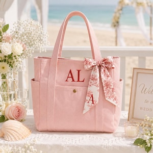 Pode incluir: Uma bolsa tote de lona rosa claro com as iniciais AL bordadas em vermelho no bolso frontal. A bolsa é acentuada com uma fita decorativa com padrão toile de Jouy branco e vermelho amarrada em laço, exibindo também uma letra A vermelha.