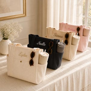 May include: Four canvas tote bags in cream, black, beige, and pink, each featuring a name embroidered in cursive script. Each bag includes a side pocket holding a tumbler with a straw and a pair of sunglasses hanging from the handle strap.