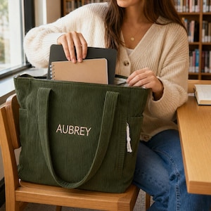 Könnte beinhalten: Eine dunkel olivgrüne Cord-Tragetasche mit dem Namen AUBREY, der in heller Garnfarbe auf der Vorderseite eingestickt ist. Die Tasche verfügt über zwei stabile Schulterriemen, Seitentaschen und einen Reißverschluss mit Notizbuch und Tablet.