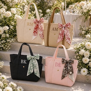 May include: Four canvas tote bags in cream, tan, black, and light pink. Each bag features two top handles, side pockets, a gold-tone snap button, and a decorative ribbon bow. The bags are personalised with embroidered initials: RM, AM, BK, and DE.