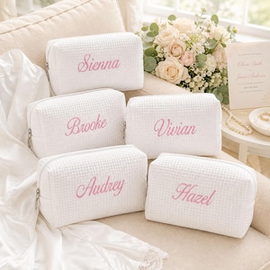 May include: Five white rectangular waffle-textured cosmetic bags with silver zippers. Each bag features a name embroidered in pink cursive script: Sienna, Brooke, Vivian, Audrey, and Hazel. The bags are arranged on a soft, light-colored surface.