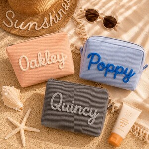 May include: Three rectangular corduroy pouches in peach, light blue, and dark gray. Each pouch features a name in raised, cursive lettering: Oakley in white, Poppy in blue, and Quincy in gray. The pouches have a zipper closure along the top.