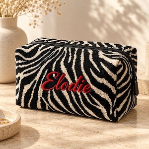 May include: A rectangular zippered pouch featuring a black and white zebra print pattern with a textured surface. The name Elodie is embroidered in red cursive script on the front center of the pouch, which has a black zipper closure.