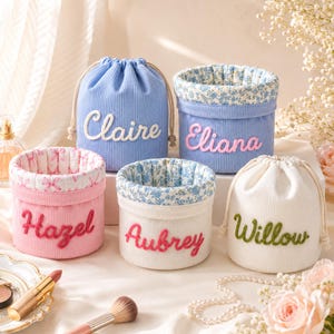 Op de afbeelding: Vijf cilindrische opbergbakjes van corduroy. Twee zijn trekkoordzakjes in lichtblauw en wit, en drie zijn open mandjes in roze, wit en blauw. Elk is voorzien van de geborduurde namen: Claire, Eliana, Hazel, Aubrey en Willow.