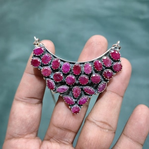 May include: A crescent-shaped pendant featuring numerous faceted, deep pinkish-red ruby gemstones set in a dark, oxidized silver metal frame. The stones are arranged in a dense, multi-layered cluster, creating a textured, geometric appearance.