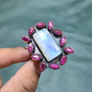 May include: A silver ring featuring a large, rectangular, iridescent moonstone cabochon at the center, surrounded by a cluster of twelve small, marquise-cut, deep pink ruby gemstones set in dark, oxidized metal prongs and bezels.