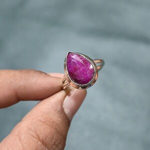 May include: A ring featuring a faceted, pear-shaped, deep pink gemstone set in a polished silver-toned metal bezel. The band is split into two thin, parallel silver-toned metal wires that connect to the base of the gemstone setting.