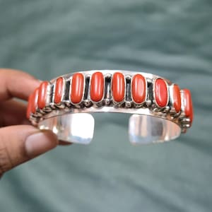 May include: A silver cuff bracelet featuring a row of eleven smooth, oval-shaped red stones set vertically. The metal band has a polished finish with small silver bead accents placed between each stone, creating a consistent, repeating pattern.