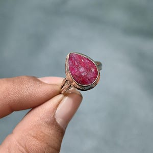 Può includere: Un anello con una grande pietra preziosa di rubino grezzo a forma di pera, con una tonalità magenta profonda e una superficie cristallina testurizzata. La pietra è incastonata in una semplice montatura in metallo argentato con una fascia sottile.