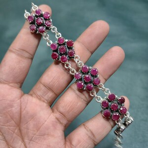 May include: A silver bracelet featuring four flower-shaped clusters, each composed of six round, deep pinkish-red faceted gemstones set in dark metal. The floral segments are connected by silver circular links, creating a continuous chain design.