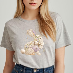 May include: A light gray t-shirt featuring a detailed embroidered bunny design. The bunny is adorned with floral accents, a large bow, and a lavender vest. Small butterflies and floral details are scattered around the bunny.