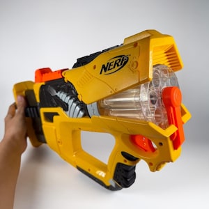 Könnte beinhalten: Ein gelb-schwarzer Nerf-Spielzeugblaster mit einer durchsichtigen, rotierenden Trommel, einem orangefarbenen Abzug und einem orangefarbenen Spannhebel. An der Seite befindet sich das schwarze Nerf-Logo. Das Design umfasst schwarze Akzente.