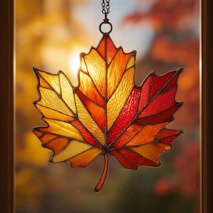 May include: A stained glass suncatcher shaped like a maple leaf, featuring segments of textured yellow, orange, and red glass held together by dark metal solder lines. The leaf hangs from a thin metal chain, catching light through its translucent panes.
