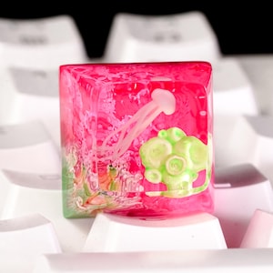 May include: A square resin mechanical keyboard keycap featuring a translucent pink base with an embedded white jellyfish figure and a cluster of light green coral-like structures. The keycap is positioned on a white mechanical keyboard.