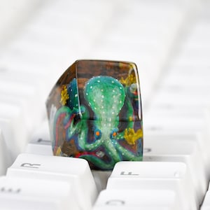 May include: A square-shaped resin keycap featuring a detailed hand-painted green octopus with orange eyes and white spots. The octopus is set against a dark, textured brown background with small yellow accents, creating a deep, layered appearance.