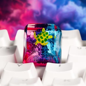 May include: A custom resin mechanical keyboard keycap featuring a yellow and green sea turtle encased within a clear, multi-colored block. The interior displays swirling shades of pink, purple, and blue, positioned on a white keyboard.