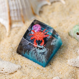 May include: A square resin mechanical keyboard keycap featuring a clear top section with a small red crab and a pink sea anemone inside. The base of the keycap transitions from a dark gray to a vibrant blue, depicting ocean waves and sea foam.