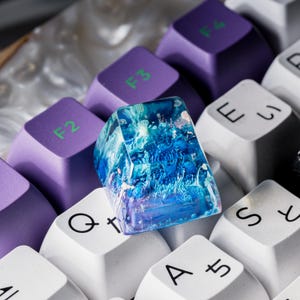 May include: A custom artisan mechanical keyboard keycap featuring a translucent blue and white resin design with swirling patterns, positioned among purple F-series keys with green text and white keys with black alphanumeric characters.