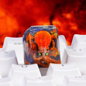 Peut inclure: Une touche de clavier mécanique personnalisée en résine représentant une pieuvre orange détaillée aux yeux verts, incrustée dans un bloc transparent et brillant. La touche est placée sur un clavier blanc, révélant une scène aquatique.