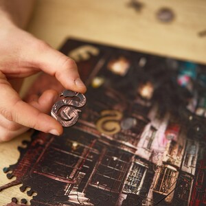 May include: A person holding a wooden puzzle piece shaped like the letter "S". The piece is brown with a textured surface. The partially assembled puzzle depicts a detailed street scene with buildings.