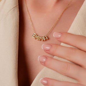 May include: A gold necklace spelling out the name "EMMA" in individual letter beads. The necklace is worn around the neck. The person is wearing a beige blazer.