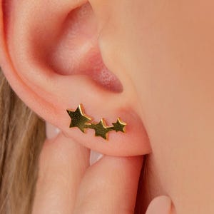May include: Gold-colored star-shaped earrings. The earrings feature three stars in a graduated design, worn on the ear. The image shows a close-up of the ear and part of the hand.