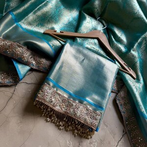 May include: A folded teal fabric with a blue border and intricate embroidery. The fabric is draped over a wooden hanger, with a matching teal fabric in the background. The embroidery features a floral design with small, dangling beads.