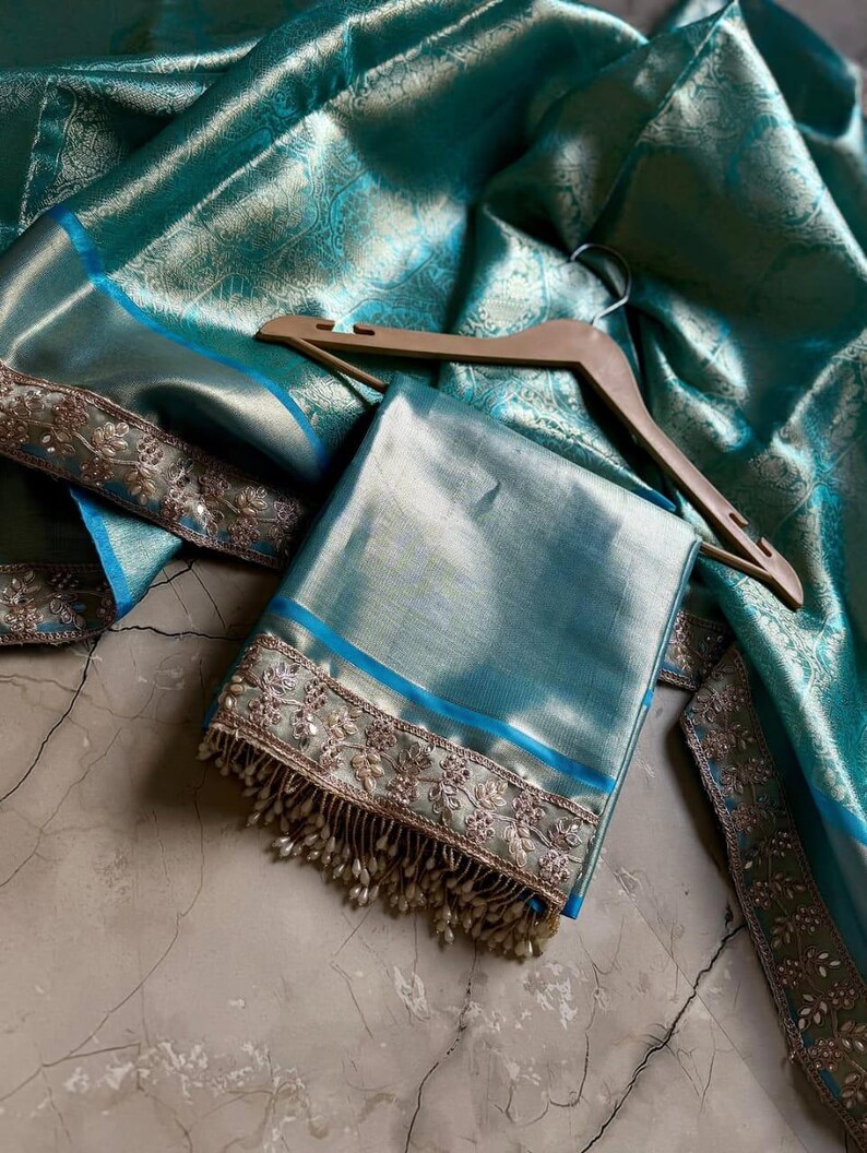 May include: A folded teal fabric with a blue border and intricate embroidery. The fabric is draped over a wooden hanger, with a matching teal fabric in the background. The embroidery features a floral design with small, dangling beads.