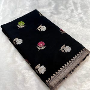 Puede incluir: Tela negra con patrones florales bordados en blanco, rosa y verde. La tela tiene un borde decorativo con un diseño detallado. El textil está doblado y se muestra sobre una superficie blanca texturizada.
