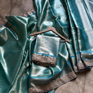May include: A teal-colored silk saree with intricate floral patterns and a shimmering finish. The saree features a silver border with detailed embroidery and a matching teal blouse piece. The fabric is draped over a wooden hanger.