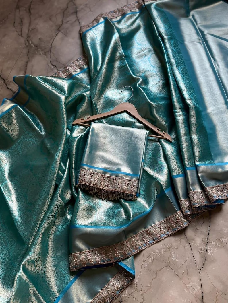May include: A teal-colored silk saree with intricate floral patterns and a shimmering finish. The saree features a silver border with detailed embroidery and a matching teal blouse piece. The fabric is draped over a wooden hanger.