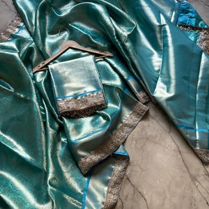 May include: A teal-colored sari with a shimmering, textured finish. The fabric is draped over a wooden hanger, with a matching square piece of fabric below. The sari features silver and gold trim along the edges and a blue border.