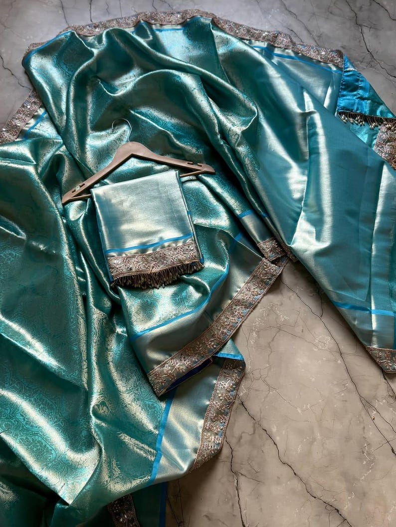 May include: A teal-colored sari with a shimmering, textured finish. The fabric is draped over a wooden hanger, with a matching square piece of fabric below. The sari features silver and gold trim along the edges and a blue border.