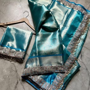 May include: A teal-colored sari with intricate silver embroidery and a matching blouse piece. The fabric has a subtle sheen and a delicate floral pattern. The sari is draped over a wooden hanger, showcasing its elegant design and texture.