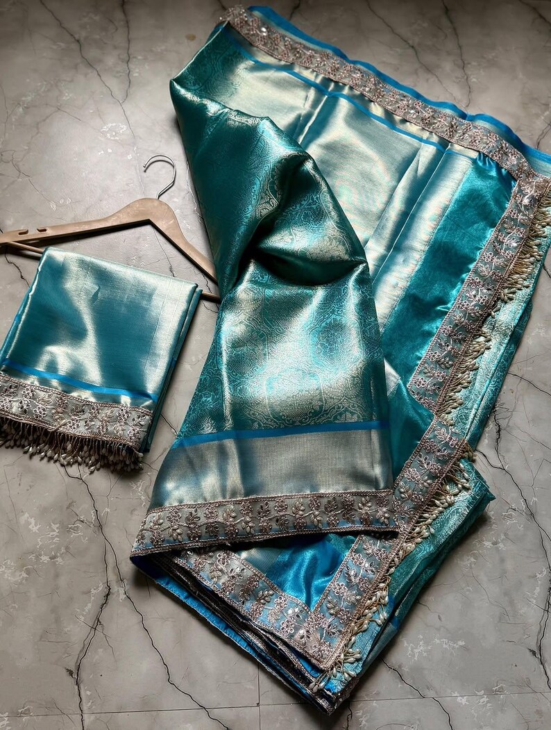 May include: A teal-colored sari with intricate silver embroidery and a matching blouse piece. The fabric has a subtle sheen and a delicate floral pattern. The sari is draped over a wooden hanger, showcasing its elegant design and texture.