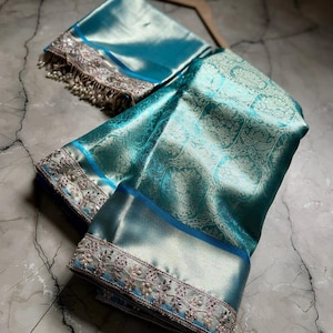 May include: A teal-colored silk saree with intricate floral patterns and a matching blouse piece. The saree features a detailed silver embroidered border with delicate beadwork. The fabric has a subtle sheen, and the ensemble is displayed on a marble surface.