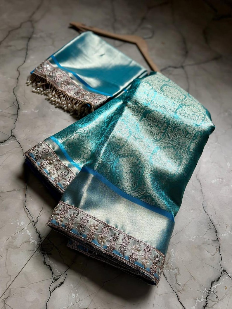 May include: A teal-colored silk saree with intricate floral patterns and a matching blouse piece. The saree features a detailed silver embroidered border with delicate beadwork. The fabric has a subtle sheen, and the ensemble is displayed on a marble surface.