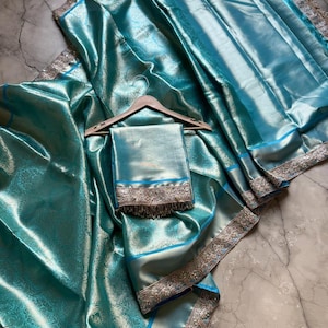 May include: A teal-colored saree with a matching shawl, draped on a surface. The saree features a shimmering, textured fabric with intricate patterns. The borders are embellished with silver and blue accents, adding a touch of elegance.