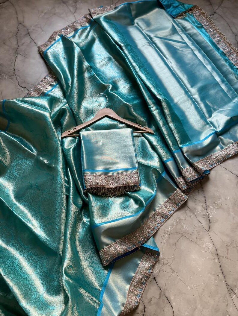 May include: A teal-colored saree with a matching shawl, draped on a surface. The saree features a shimmering, textured fabric with intricate patterns. The borders are embellished with silver and blue accents, adding a touch of elegance.