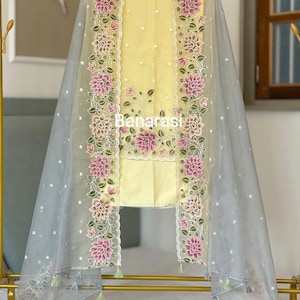 May include: A yellow salwar kameez set with floral embroidery and a sheer gray dupatta. The dupatta and the edges of the kameez feature pink and green floral designs. The word "Banarasi" is printed on the kameez.