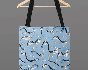 May include: A light blue tote bag featuring a pattern of black and white wavy lines and small yellow fish. The bag has black straps and is suspended from a wooden hanger.
