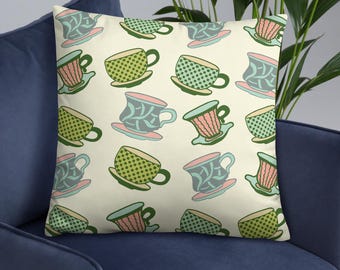 May include: Decorative throw pillow with a repeating pattern of colorful teacups. The teacups feature green polka dots, pink stripes, and blue floral designs. The pillow is on a blue couch.