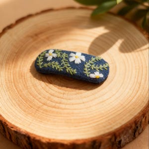 Puede incluir: Pinza para el pelo de tela azul con margaritas blancas y hojas verdes bordadas. La pinza ovalada descansa sobre una superficie de madera, mostrando un diseño floral artesanal.