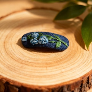 Peut inclure: Une barrette brodée bleu marine avec un motif floral. La barrette présente trois groupes de fleurs bleues avec des feuilles et des tiges vertes. Elle est posée sur une surface en bois avec un grain naturel.