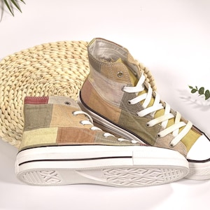 Può includere: Sneakers alte con un design patchwork nei toni del beige, marrone e verde oliva. Le scarpe hanno lacci bianchi, suola in gomma bianca e una striscia nera. Le sneakers sono appoggiate su un tappetino intrecciato.