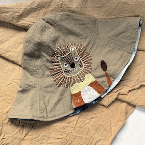 Può includere: Un cappello a secchio beige con un motivo a leone ricamato. Il leone ha una criniera marrone, una faccia beige e una coda marrone. Il cappello ha una fodera a fantasia blu e bianca ed è realizzato in un tessuto morbido.
