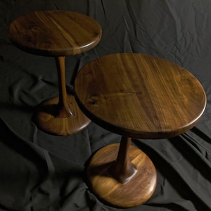 Puede incluir: Dos mesas auxiliares de madera hechas a mano con acabado en nogal oscuro. Cada mesa presenta una tapa redonda, un tallo delgado y curvado y una base circular. Las mesas se muestran sobre un fondo de tela gris oscuro.